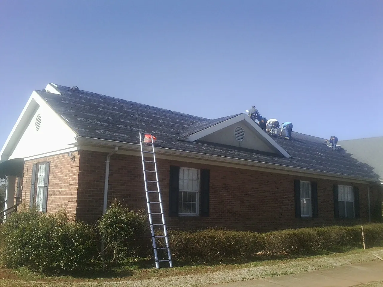 Crew tearing shingles off a brick ranch roof with a ladder set — Metal Roof Repair work by our crew in Hilton