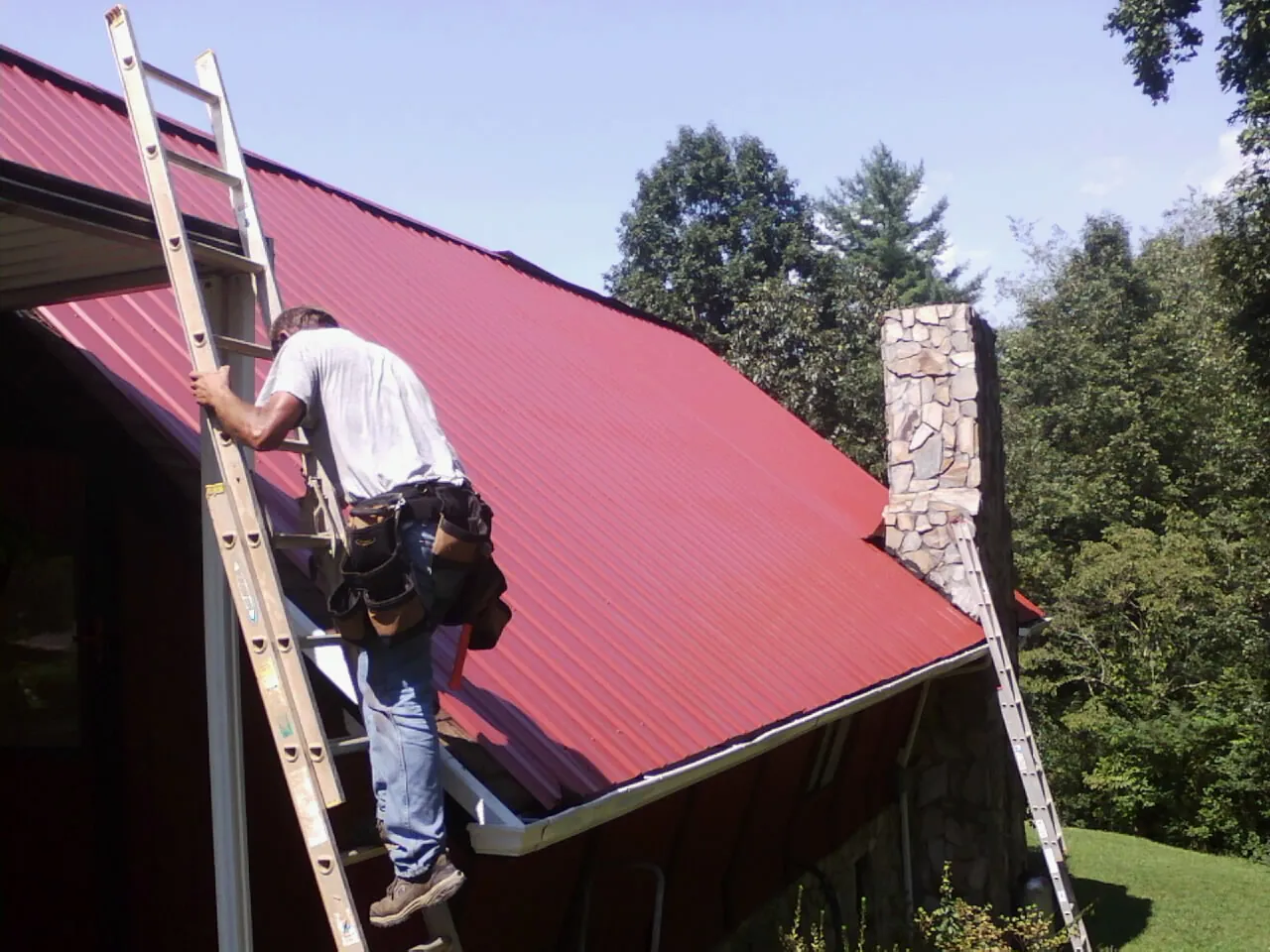 Roofer on a ladder beside a red metal roof and stone chimney — Soffit & Fascia Repair by Roofing Brothers in Hilton